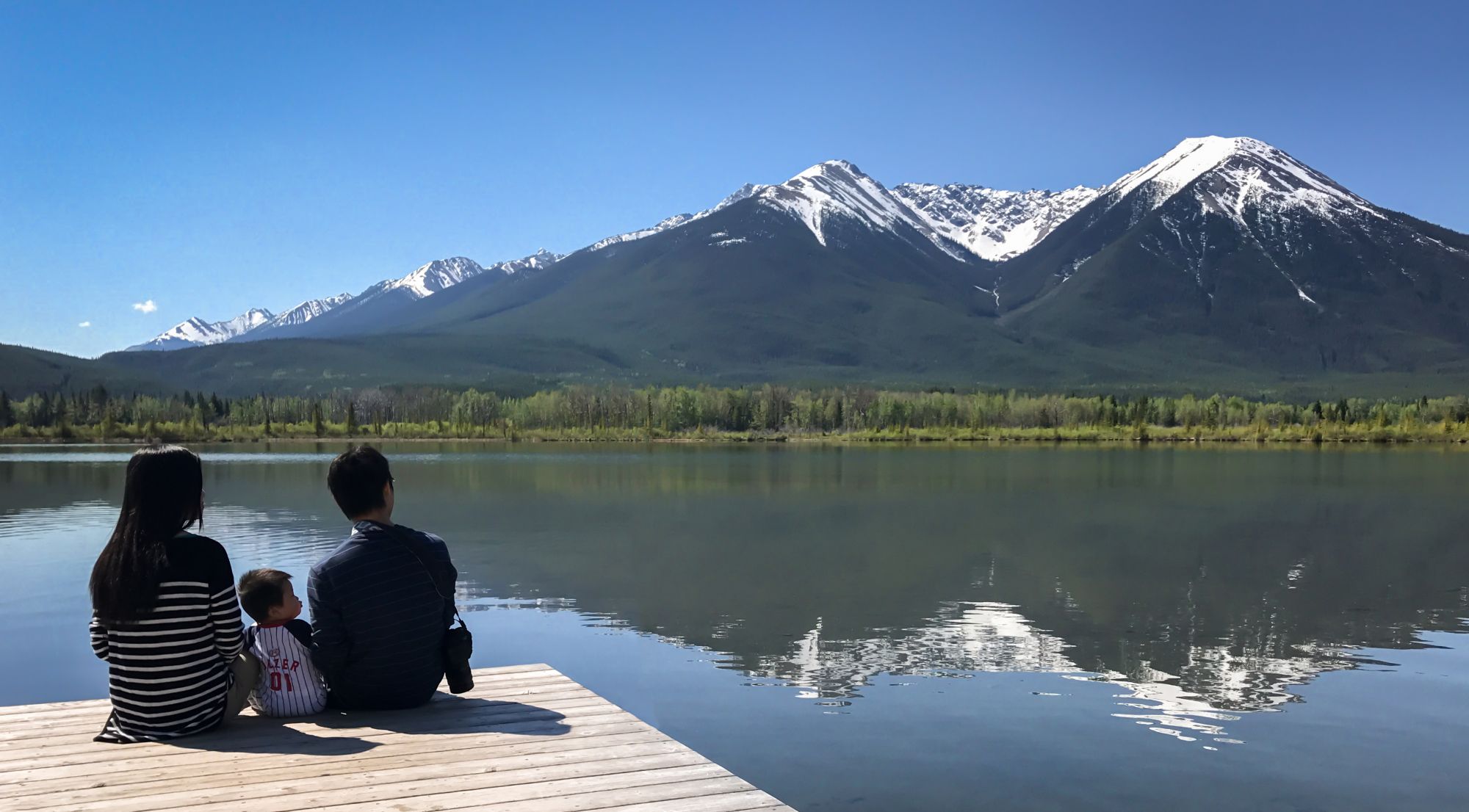 A young family sitting on a wooden pier by Vermilion Lakes, with the Rocky Mountains behind them